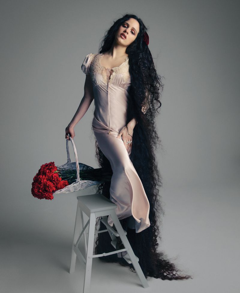 Rosalía posing in a blush satin gown with floor-length dark wavy hair, holding a basket of red flowers on a white stool.