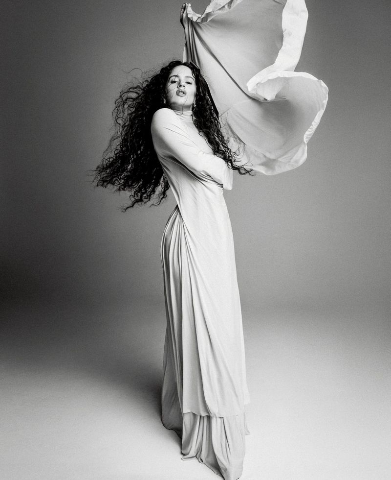 Rosalía poses in a draped long-sleeve dress with windblown hair and billowing fabric against a studio backdrop in black and white.