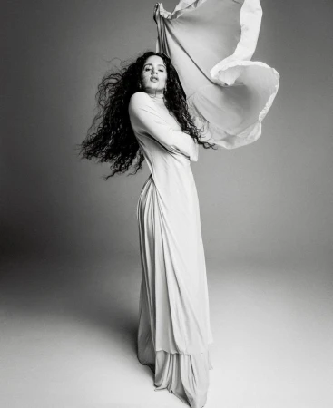 Rosalía poses in a draped long-sleeve dress with windblown hair and billowing fabric against a studio backdrop in black and white.