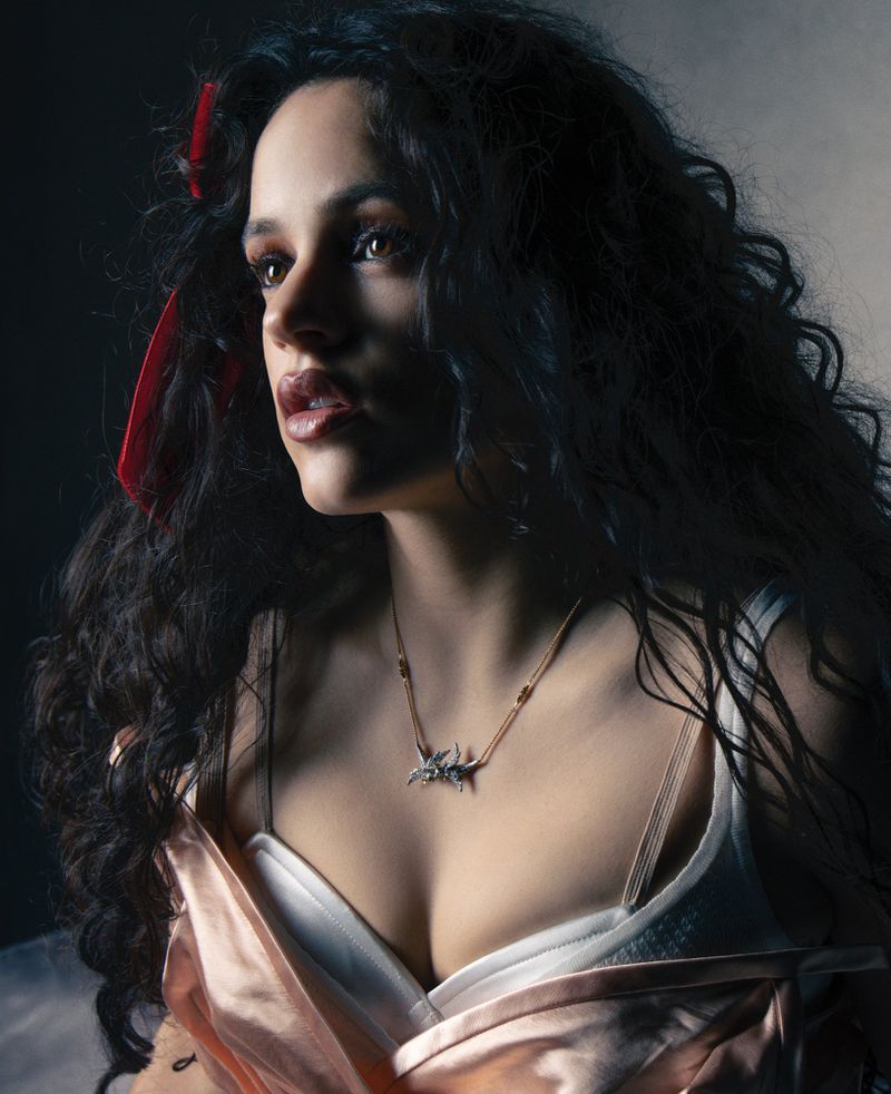 Portrait of a woman with dark curly hair and a red ribbon. She wears a light pink dress and a statement necklace in a dimly lit environment.