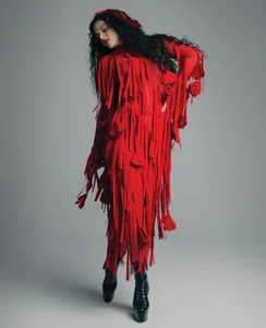 Woman with long dark hair wearing a red fringed dress and platform boots against a plain grey backdrop.