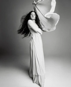 Rosalía poses in a draped long-sleeve dress with windblown hair and billowing fabric against a studio backdrop in black and white.