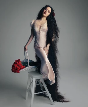Rosalía posing in a blush satin gown with floor-length dark wavy hair, holding a basket of red flowers on a white stool.