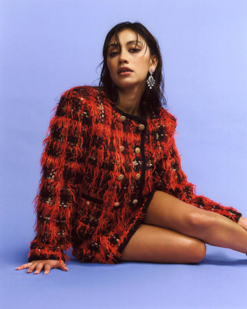 Woman in a red tweed jacket with pearl buttons sits against a lilac background, showcasing fashion, style, and portrait photography.