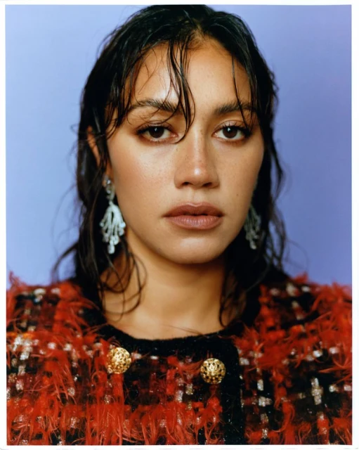 Close-up portrait of a young adult female with wet dark hair and light skin, wearing a red and black tweed jacket against a blue background.