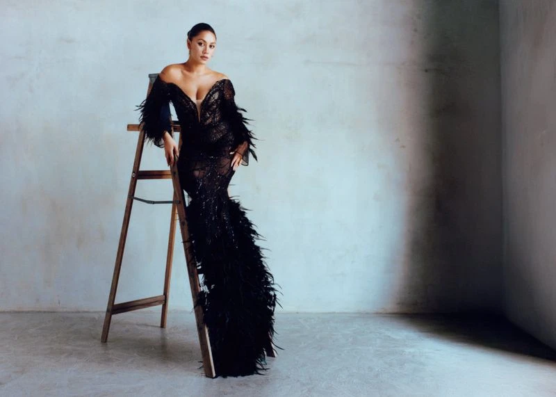 Adult woman in black feathered gown poses on a ladder indoors against a neutral wall, captured in a full-length shot with soft lighting.