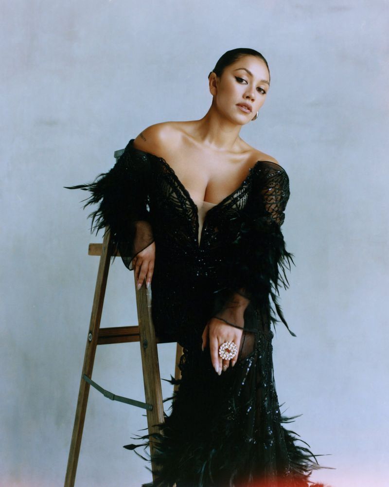 A woman poses indoors, seated on a wooden ladder; she wears a black, feathered, formal dress and large ring, against a neutral background.
