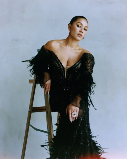 A woman poses indoors, seated on a wooden ladder; she wears a black, feathered, formal dress and large ring, against a neutral background.