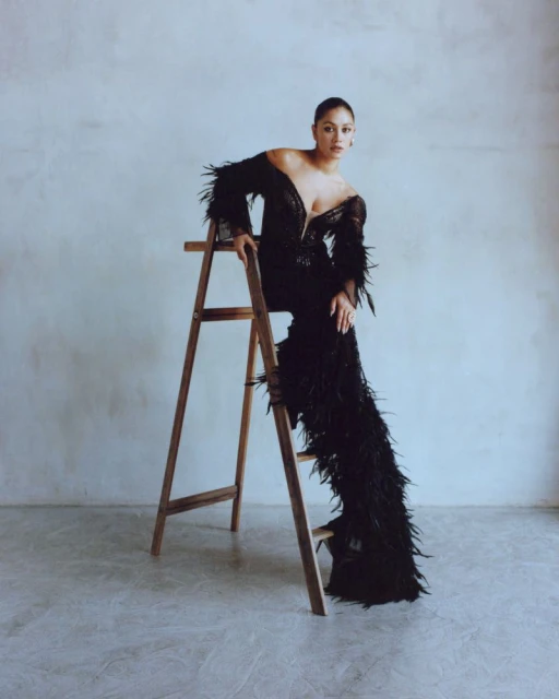 Woman in a black feather dress sits on a wooden ladder in front of a pale wall, showcasing fashion and beauty in a studio setting.