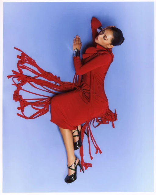 Woman in a red dress and black high heels lying on her side against a light purple background in a studio shot.
