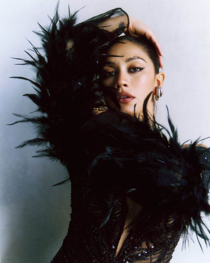 Glamorous portrait of a woman with black feathered boa. She is wearing jewelry and posing with her hand on her head.