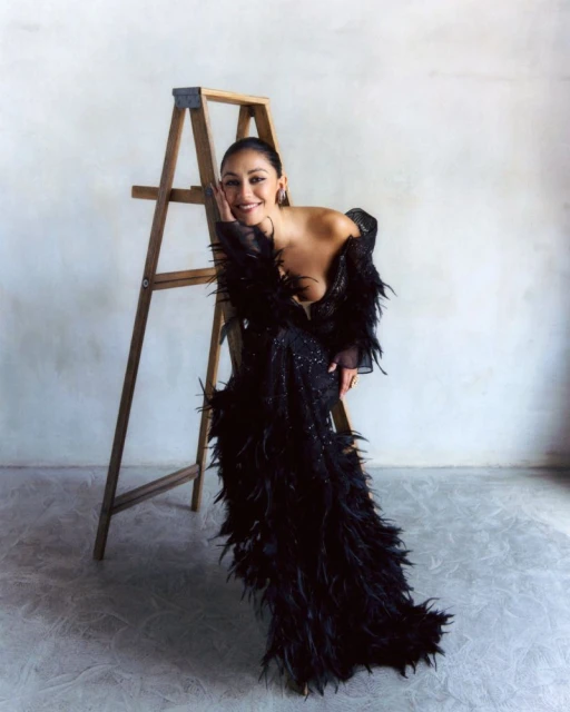 Woman poses in a black feather gown, leaning on a ladder in front of a white wall and grey floor. Soft lighting and elegant fashion.