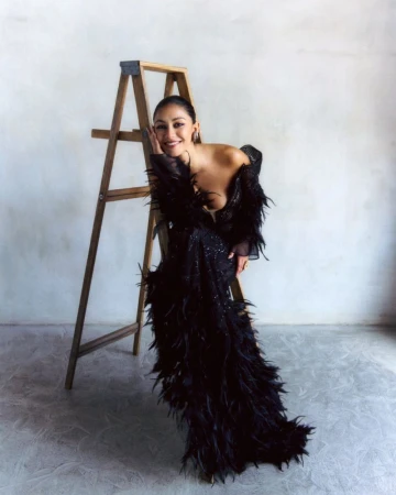 Woman poses in a black feather gown, leaning on a ladder in front of a white wall and grey floor. Soft lighting and elegant fashion.