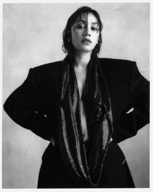 Monochrome portrait of a woman with wet hair, wearing an oversized blazer and shimmering scarf, hands on hips against a grey backdrop.
