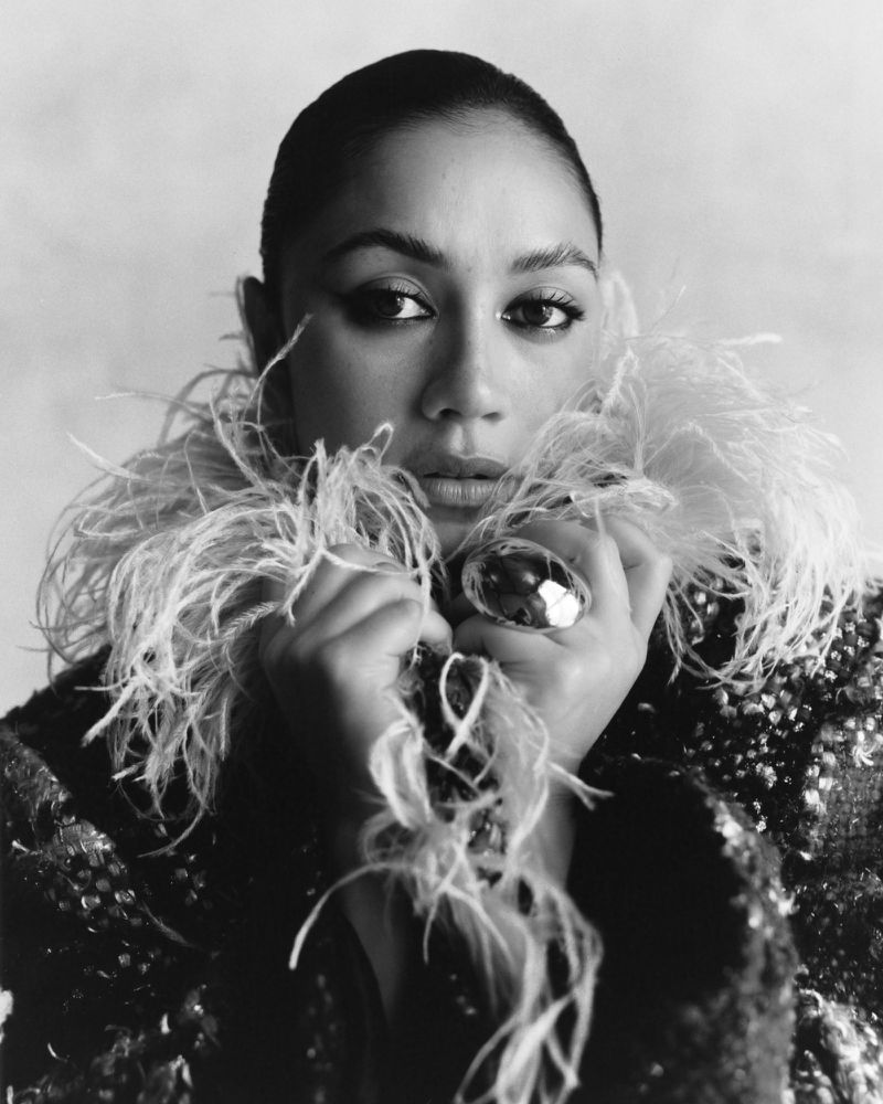 Headshot of a person with hands near their face, wearing a large ring and a feathered collar, in a black and white portrait against a light background.