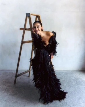 Luciane Buchanan smiling in a dramatic black feather gown while seated on a wooden ladder against a neutral studio backdrop.