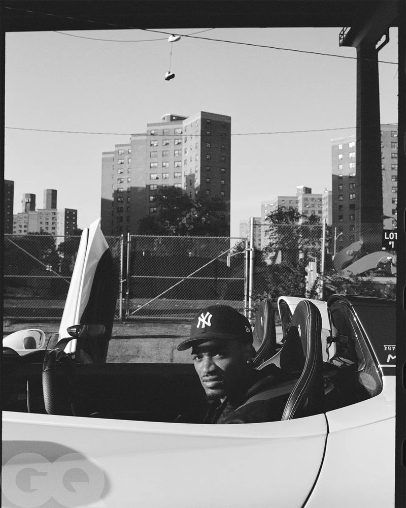 Monochrome portrait of a smiling man in a convertible car with city buildings in the background, wearing a baseball cap.