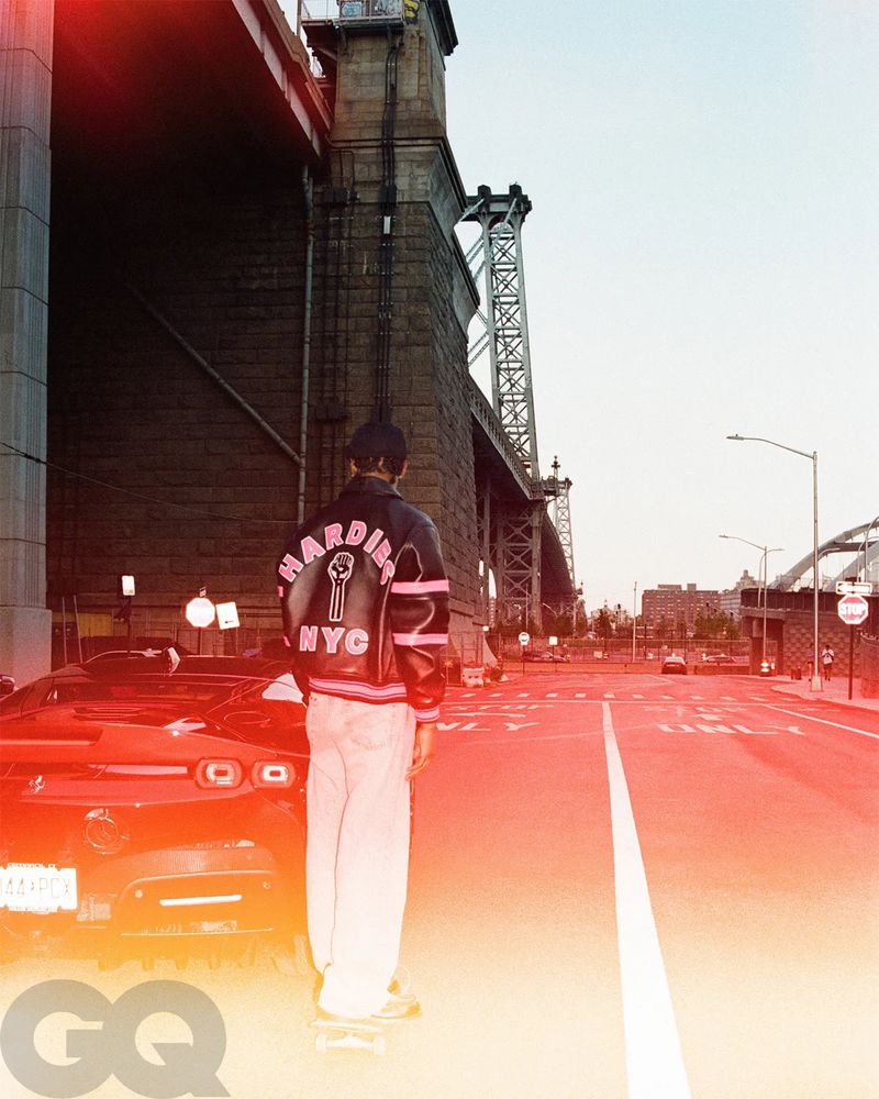 Person in jacket stands on street under bridge near car, with GQ logo in corner. Urban scene, low angle, fashion photography.