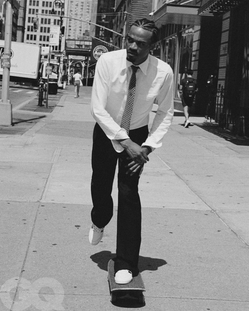 Black and white shot of a Black man in business attire skateboarding on a sidewalk in an urban setting, wearing white sneakers and a tie.