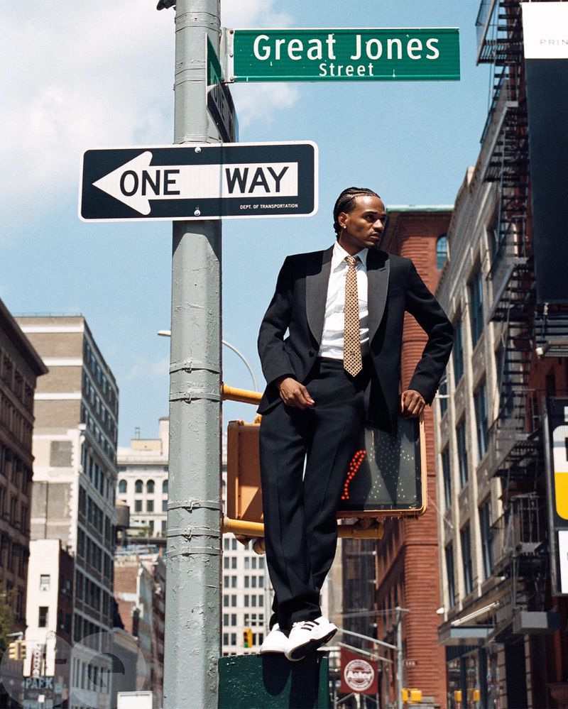 An adult male in a tuxedo poses sitting on a traffic light with city buildings and a street sign that reads 'Great Jones Street' in the background.