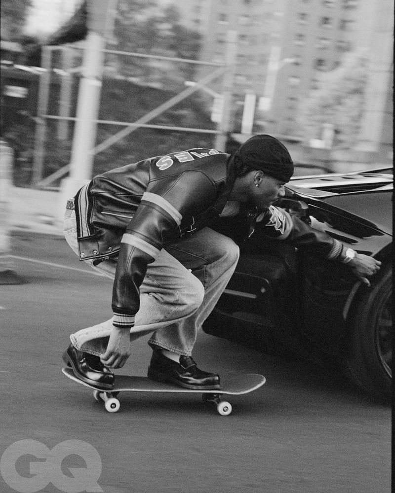 Tyshawn Jones skateboarding crouched low while holding onto a dark sports car in a black and white editorial photograph.