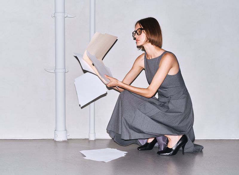 Woman in a gray dress squats on the floor holding papers and a file folder in a white indoor studio environment with pipes in the background.