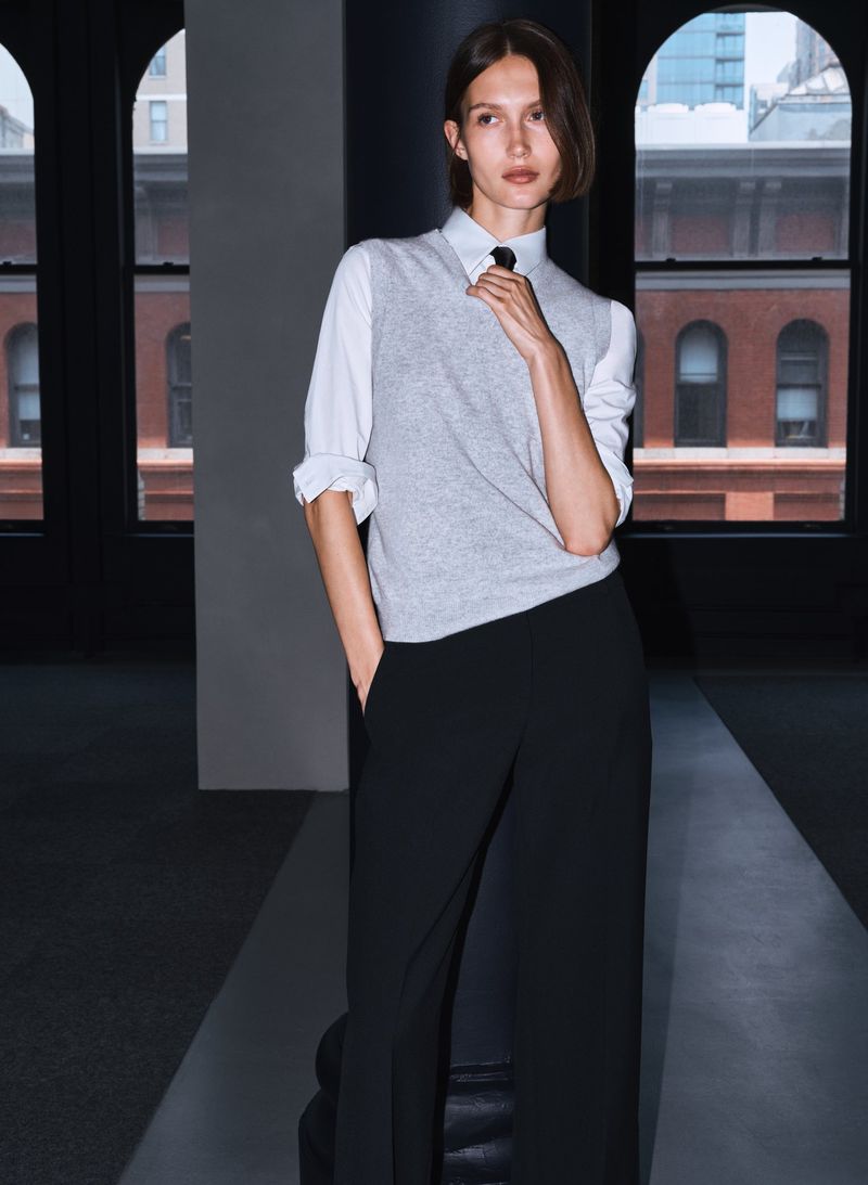Elegant woman in gray sweater vest, white shirt, and black pants stands in a naturally lit, modern interior with large windows.