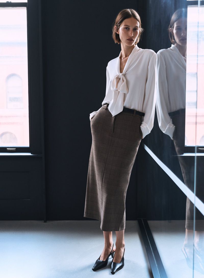 A poised fashion model in a white blouse and brown skirt stands indoors near a reflective surface and window with natural light.