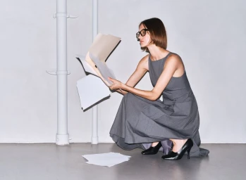 A woman in a grey sleeveless dress and glasses crouches while holding a folder and scattered papers against a neutral studio wall.