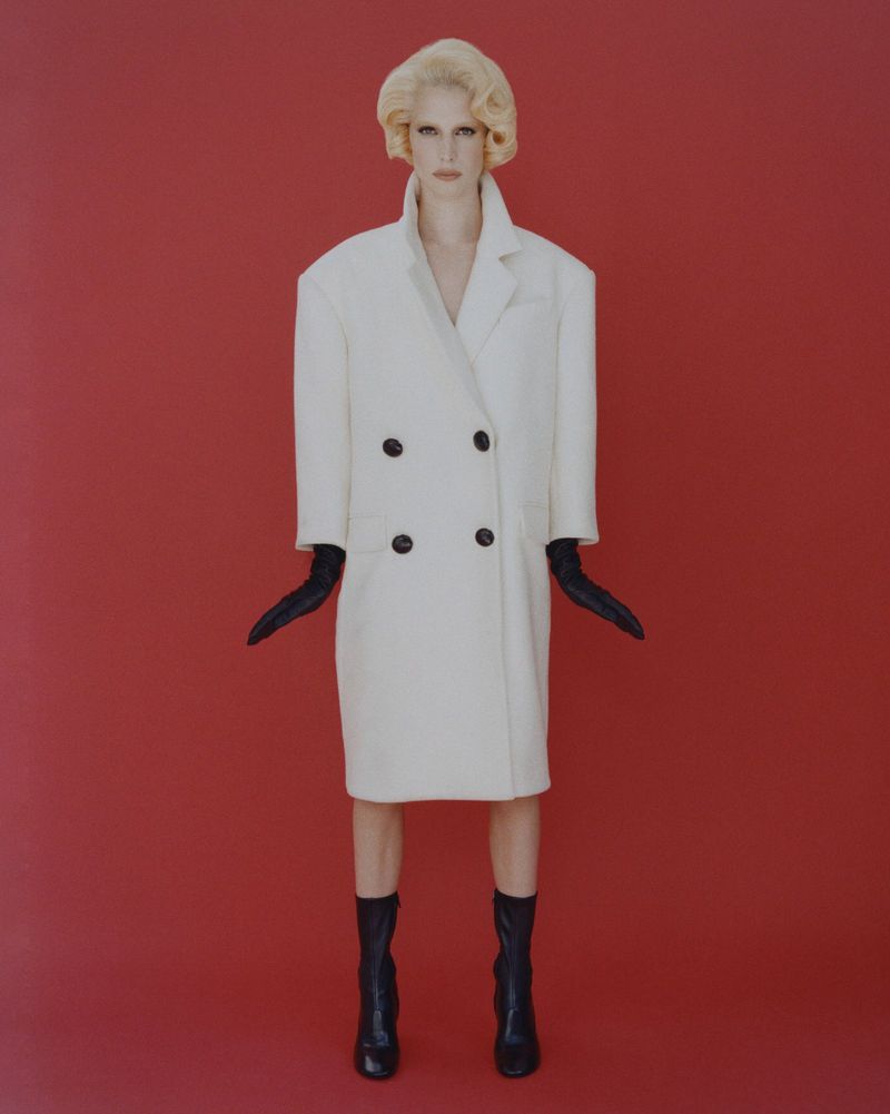 A full shot of a woman with blonde hair, wearing a white oversized coat, black gloves, and black boots on a red background.