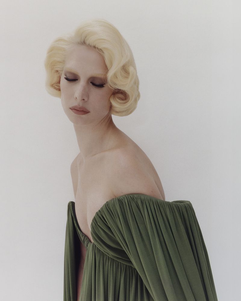 Portrait of a blond female model wearing an off-the-shoulder green dress against a white backdrop, eyes closed, in a studio setting.
