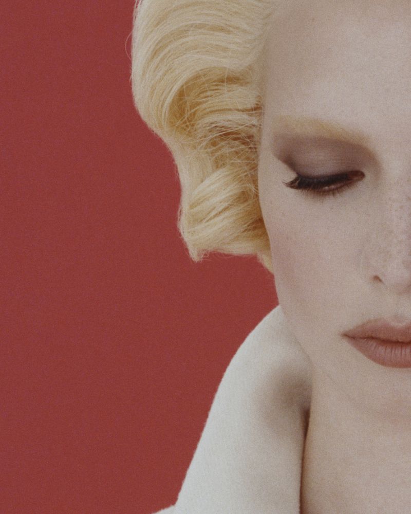 Close-up of a fair-skinned adult female model with light blonde wavy hair, dark brown eyeshadow and light red lipstick against a red backdrop.