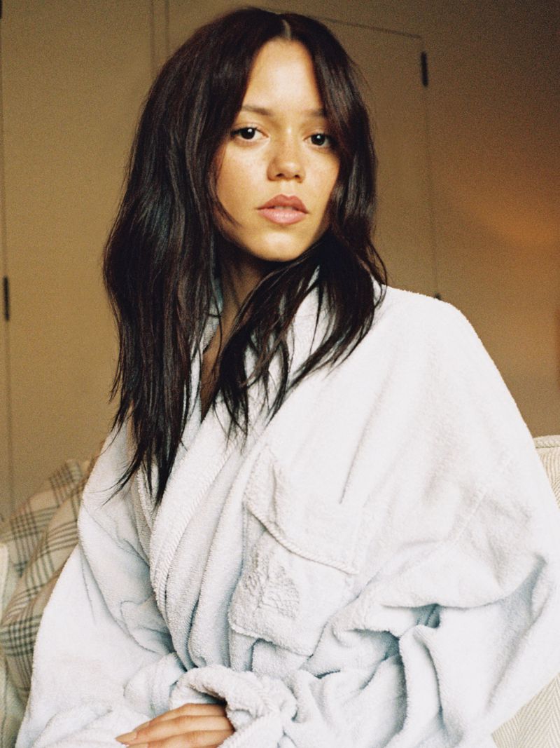 Close-up portrait of a young adult woman with brown hair wearing a white bathrobe, gazing directly at the camera in an indoor setting.