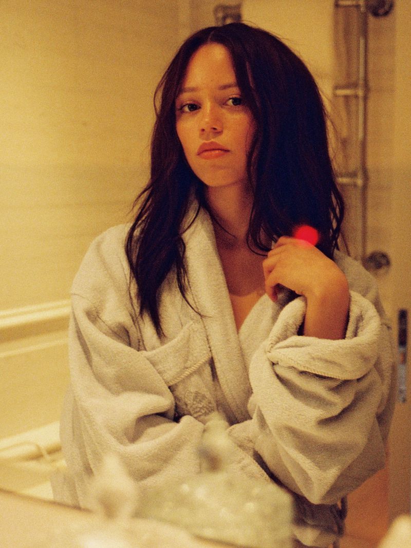 Close-up of an adult woman with dark hair in a white robe, looking at the camera in a bathroom with a mirror and neutral tones.