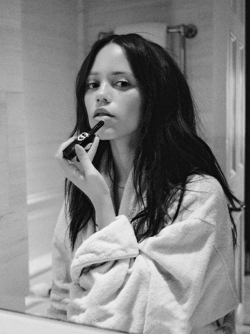 Black and white portrait of a woman in a robe applying lipstick while looking in a mirror in a bathroom.