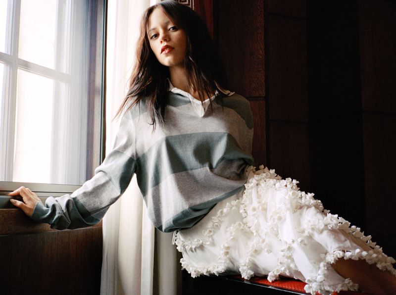 A woman poses indoors by a window in a striped collared shirt and textured skirt, leaning on the sill in soft natural light.
