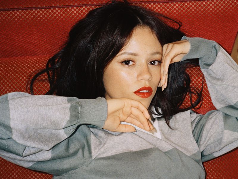 Portrait of an adult woman with dark hair and bright red lipstick lying on an orange patterned couch, resting her hands near her face.