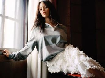 Jenna Ortega wearing a grey and green striped rugby polo and a white textured ruffled skirt while leaning against a large window.