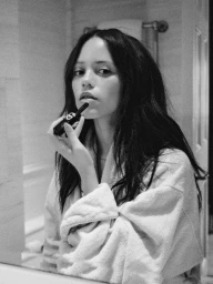 Black and white photo of Jenna Ortega in a terrycloth bathrobe applying lipstick while looking into a bathroom mirror.