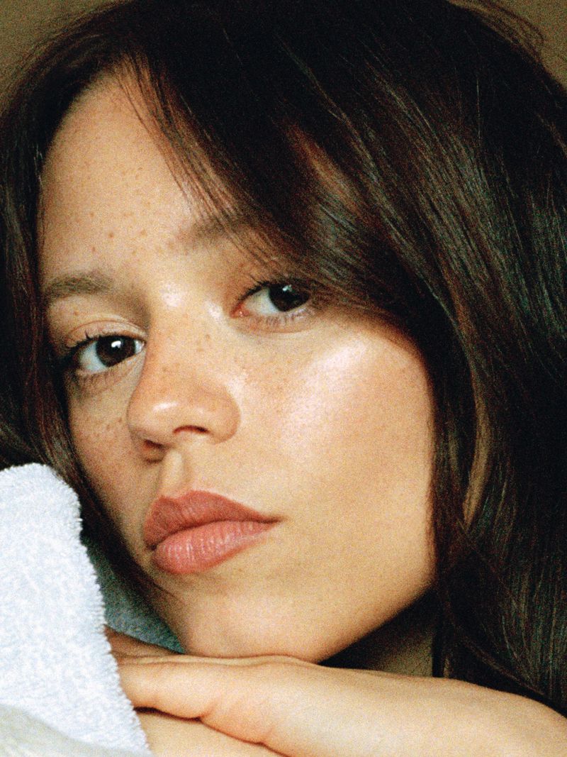 Close-up portrait of actress Jenna Ortega with dark hair, natural skin texture, and soft makeup resting chin on a white towel.