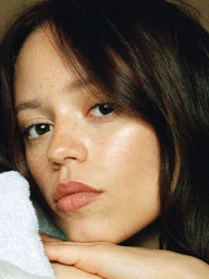 Close-up portrait of actress Jenna Ortega with dark hair, natural skin texture, and soft makeup resting chin on a white towel.