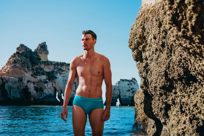 Adult male in swimwear poses on an ocean beach with blue sky, water, and rock formations in a lifestyle fashion photography setting.