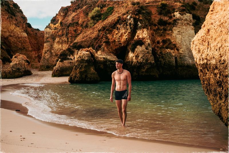 Shirtless adult male walks on a beach with rock formations and turquoise water on a sunny day, showcasing a fitness lifestyle.