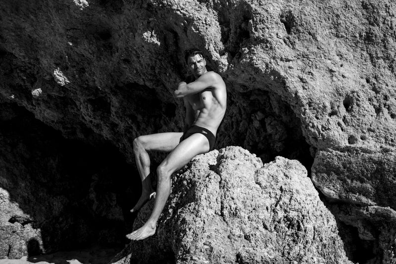 Monochrome image of a shirtless adult male sitting on a large rock formation. The man is in swimwear, set against a rocky background.