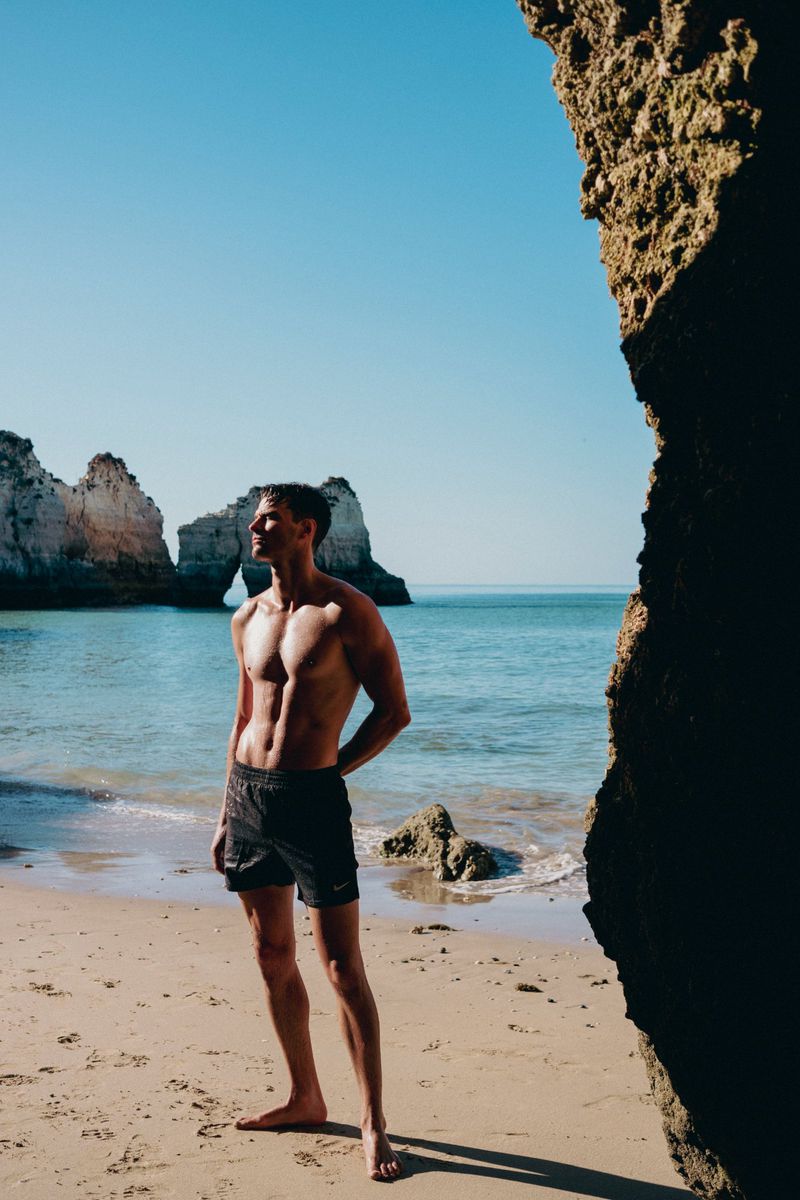 Athletic man standing on a sandy beach near a rocky coastline, under a clear blue sky, illuminated by bright sunlight.