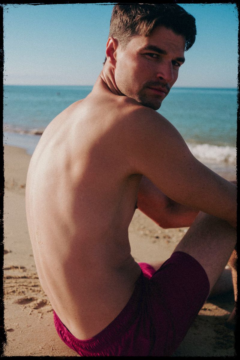 Man sitting on a sandy beach looks back over his shoulder towards the camera with the ocean and blue sky in the background.