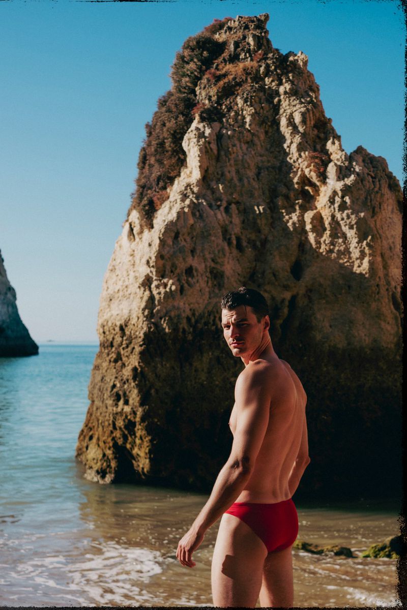 Adult male in red swimsuit poses on a beach with rock formations and blue ocean backdrop on a clear sunny day.