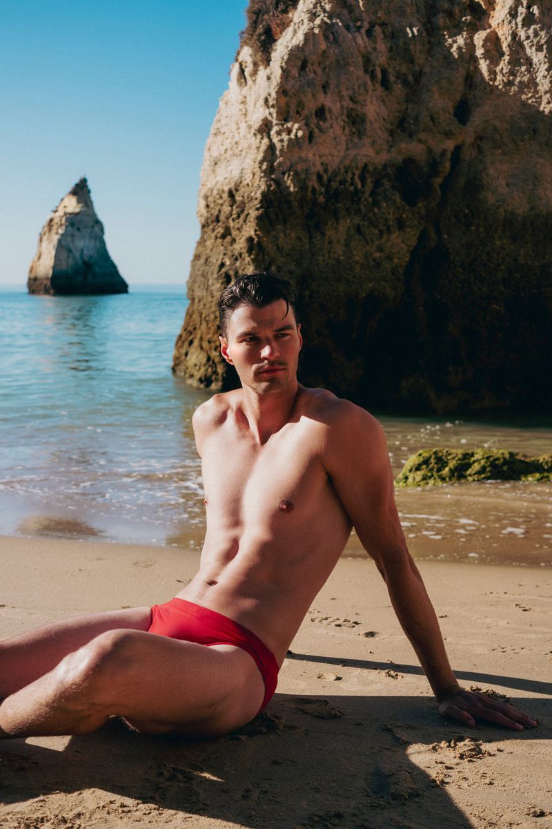 Man poses on sandy beach in red swimsuit, rocks and ocean in background, sunny day, clear sky, close-up shot of torso and face.
