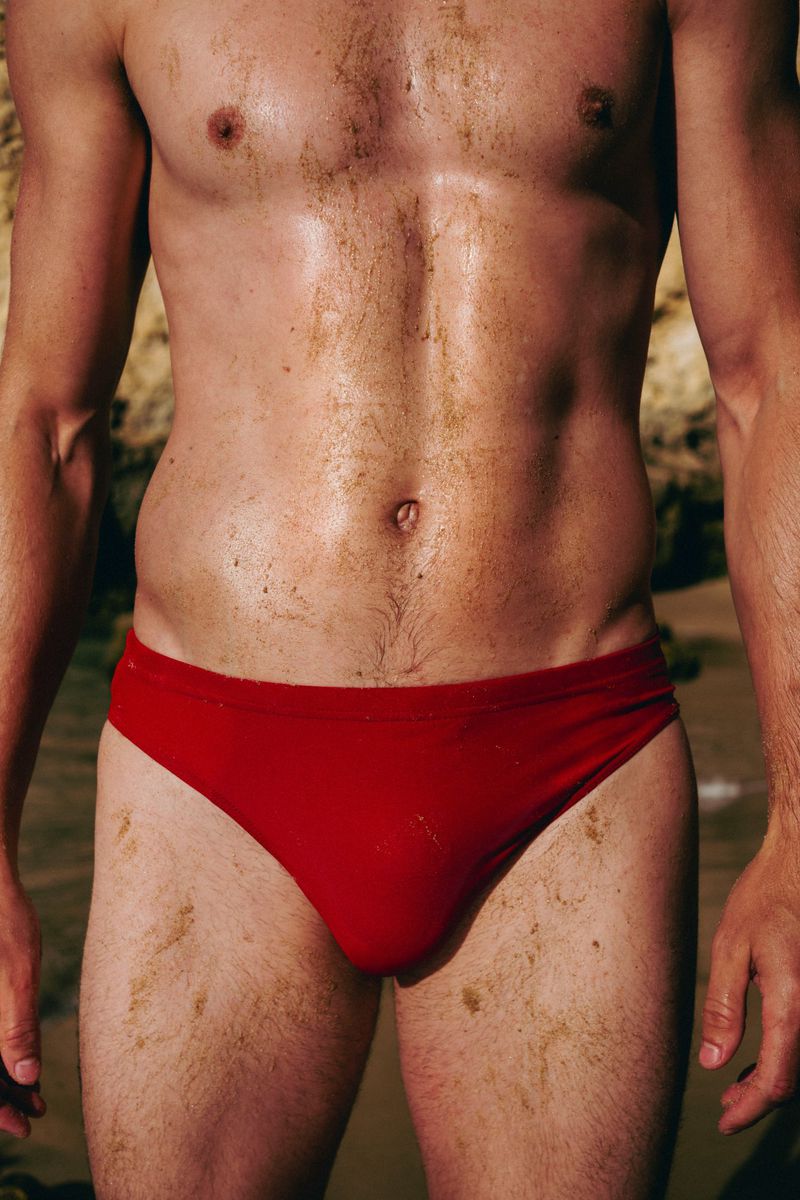 Torso of a man with an athletic build wearing red swimwear, standing on a sandy beach, with a rocky shoreline in the background.