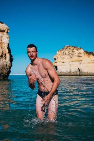 Shirtless man in black swimsuit emerges from the turquoise water in a coastal landscape featuring tan rock formations against a clear blue sky.